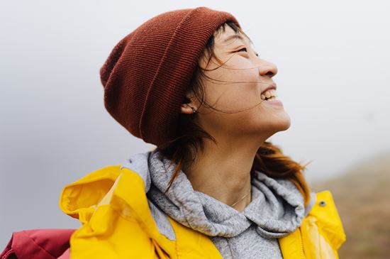 A woman in a yellow rain jacket and red beanie stands on a rainy hillside while she looks up and smiles.