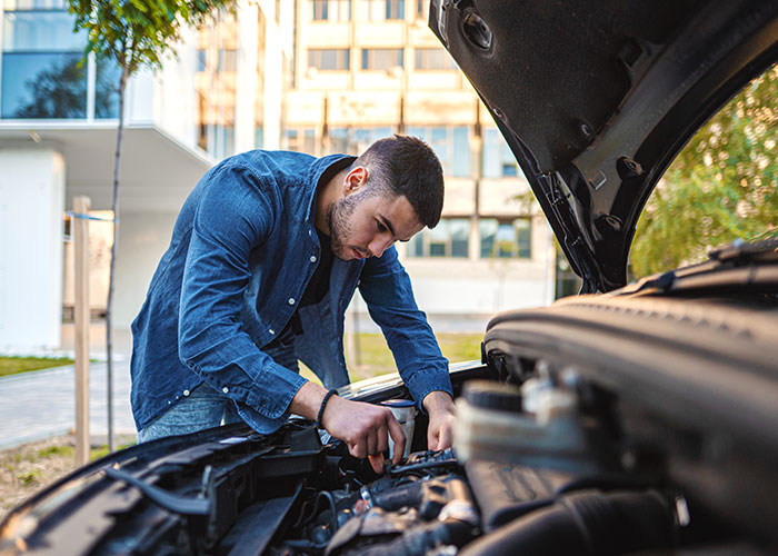 A man in a blue shirt inspects a car engine outside on the street, looking for any signs of damage.