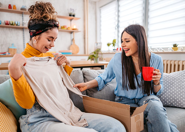 Two young women site together on a couch, one of them is holding up a shirt unpacked from a cardboard box to show the other.