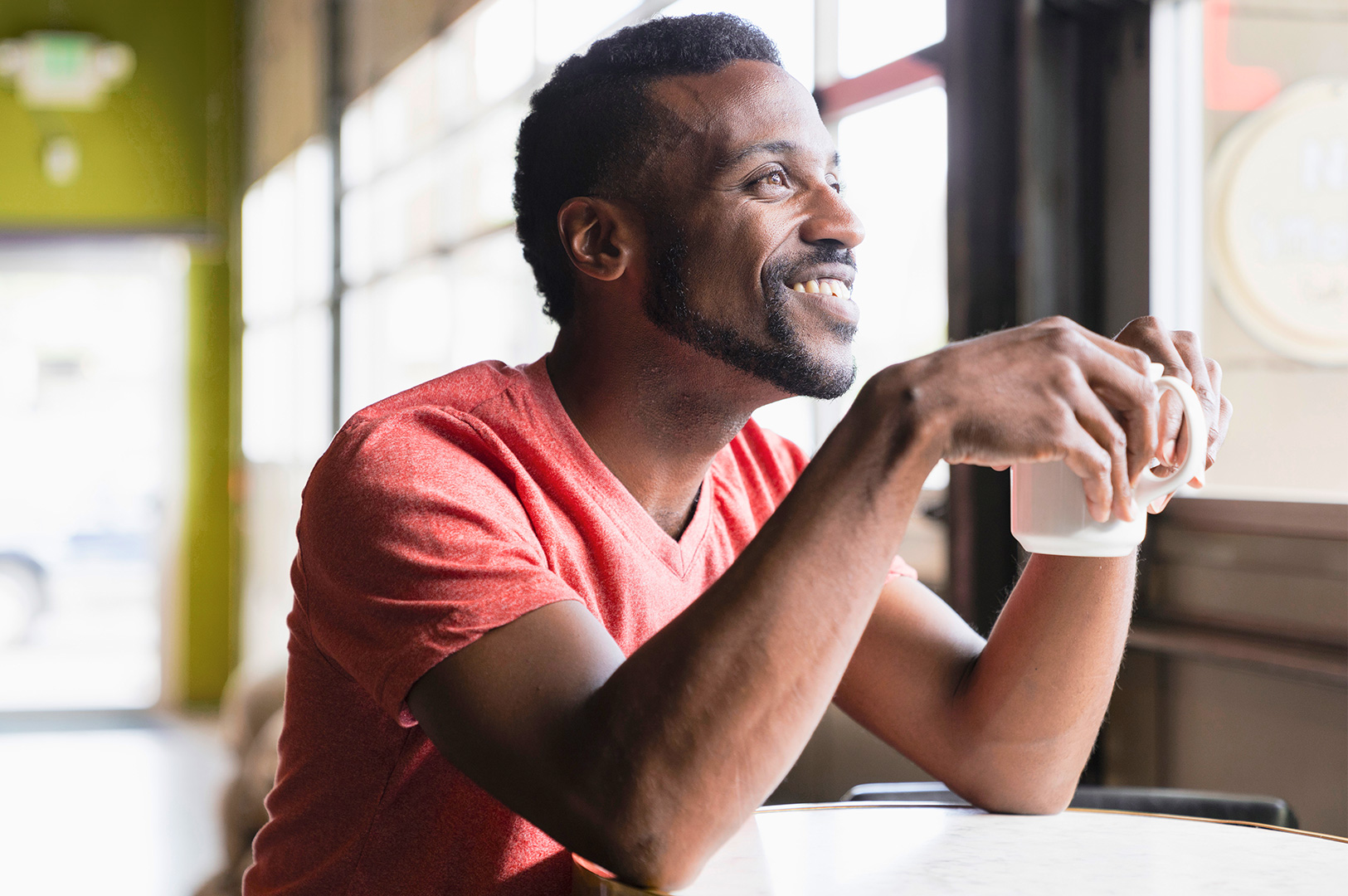 A smiling man in an orange t-shirt sits at a cafe table holding a cup of coffee. A smiling man in an orange t-shirt sits at a cafe table holding a cup of coffee.