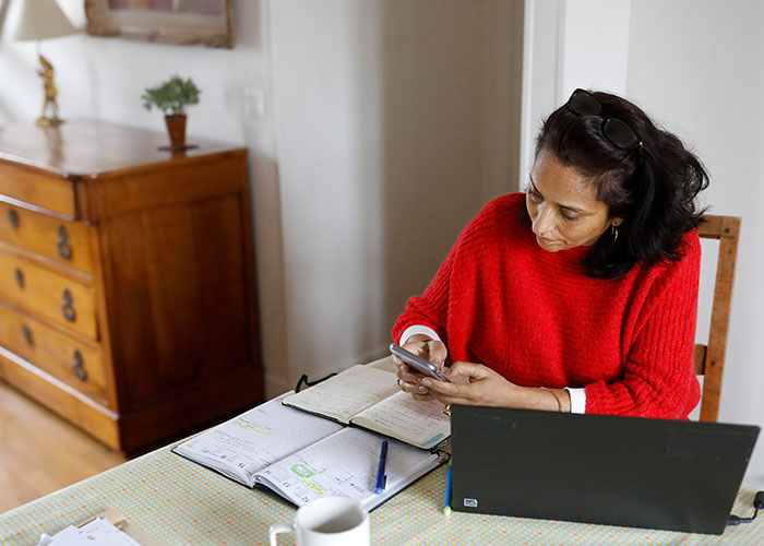 A woman sits at a table using a phone, laptop, and notebook to plan her finances