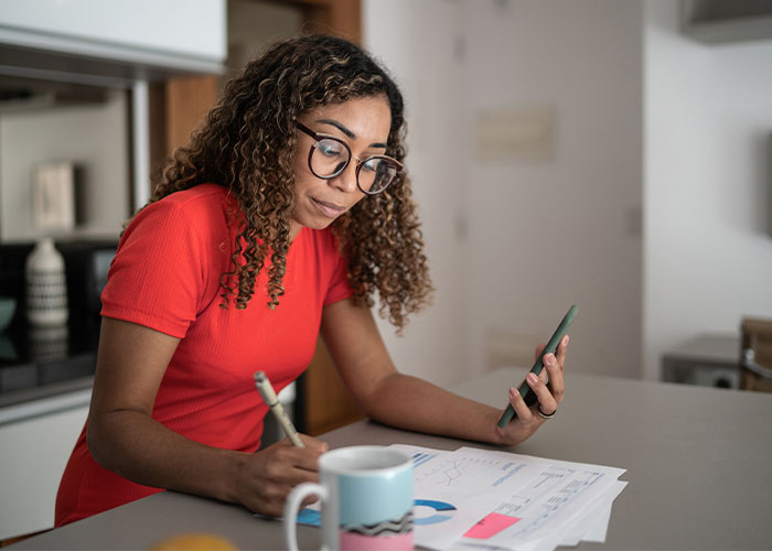 A woman sits at her kitchen table and makes notes on sheets of paper while holding a smartphone in her other hand