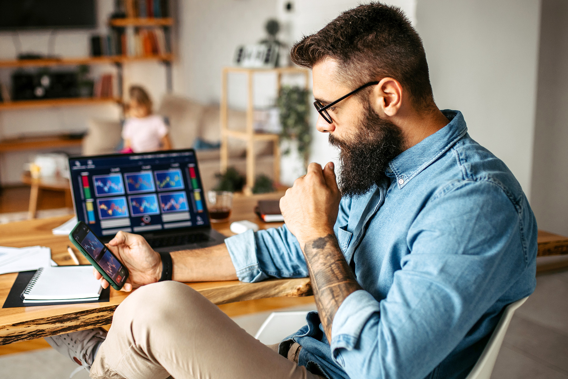 A bearded man in a blue shirt and beige pants is seated at a wooden desk, engrossed in their smartphone and laptop which display various financial graphs. 