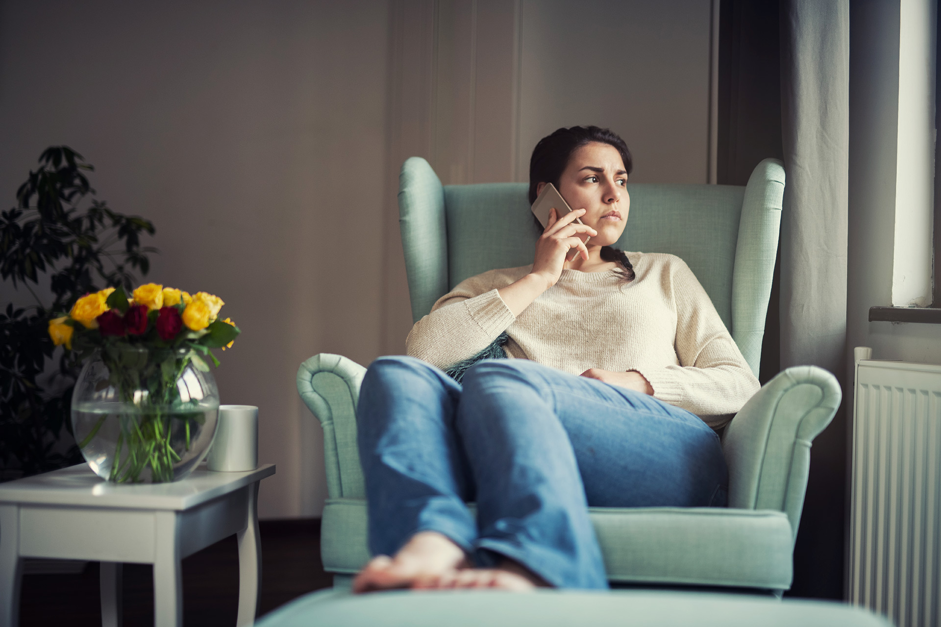 A women sits in a chair looking out of the window with a concerned look while holding a phone to her ear