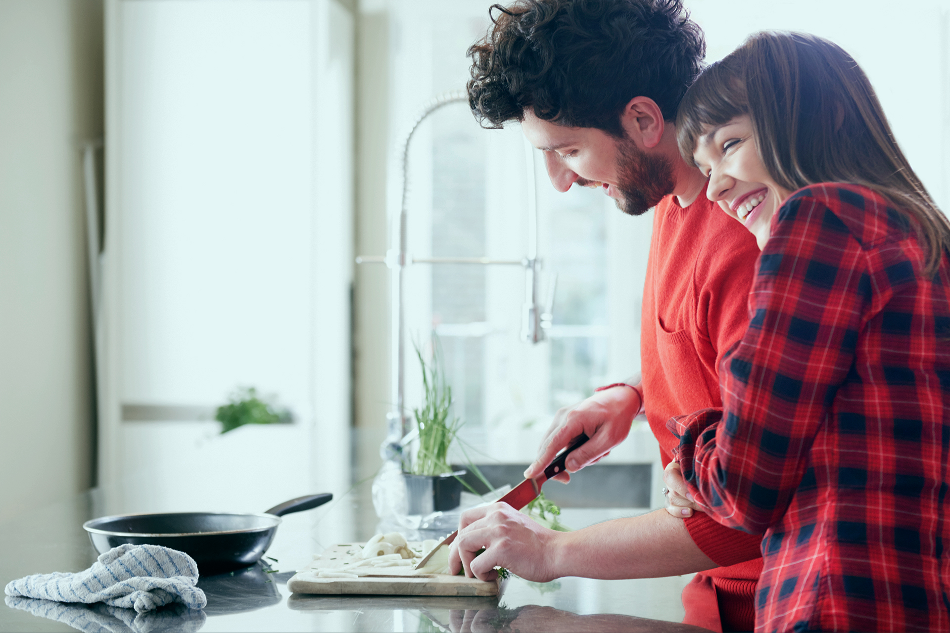 A couple is in a kitchen, a man slices vegetables on a cutting board while a woman hugs his side and smiles